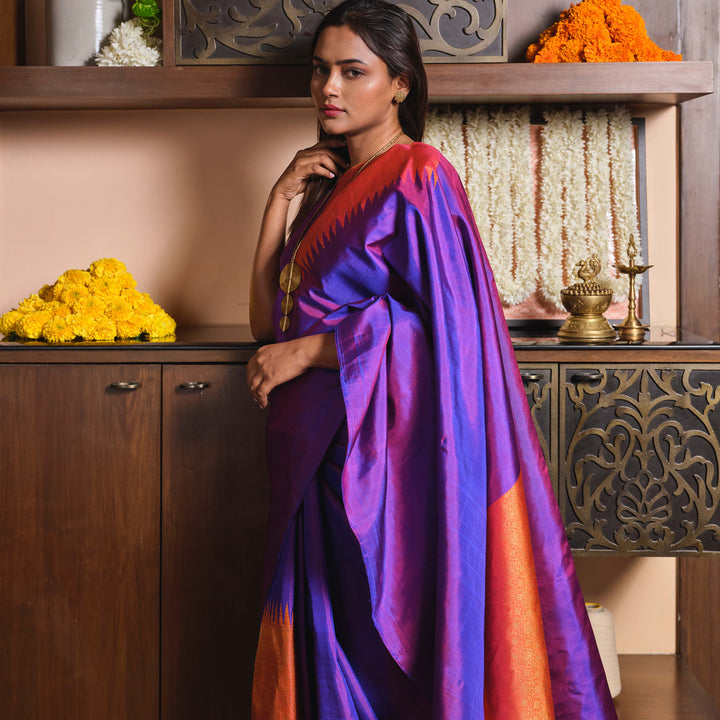 Woman in a purple saree with orange accents standing in front of decorative shelves.
