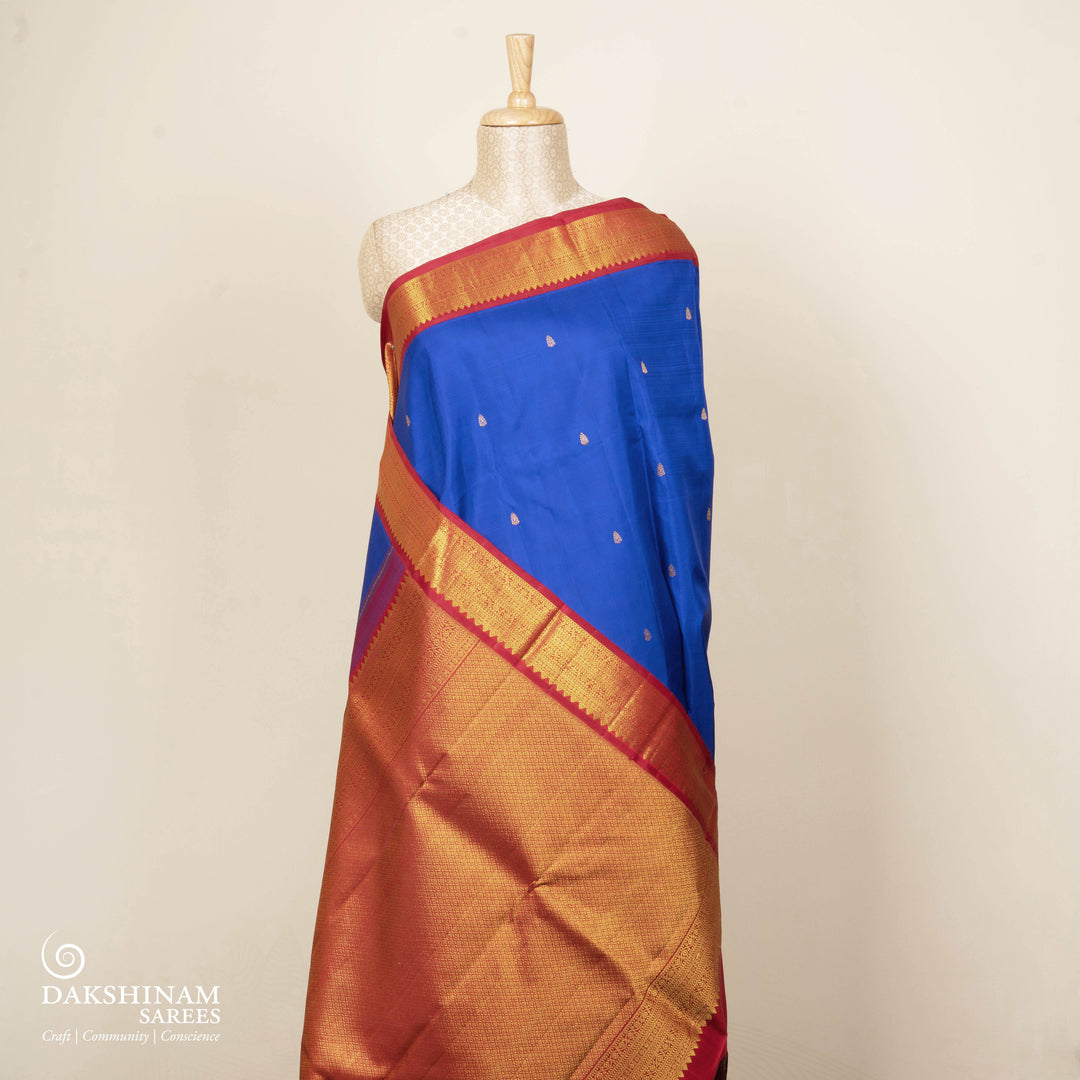 Blue handwoven Kanjivaram silk saree with gold buttas, red border with chevron and floral designs, and matching pallu, paired with plain red blouse.