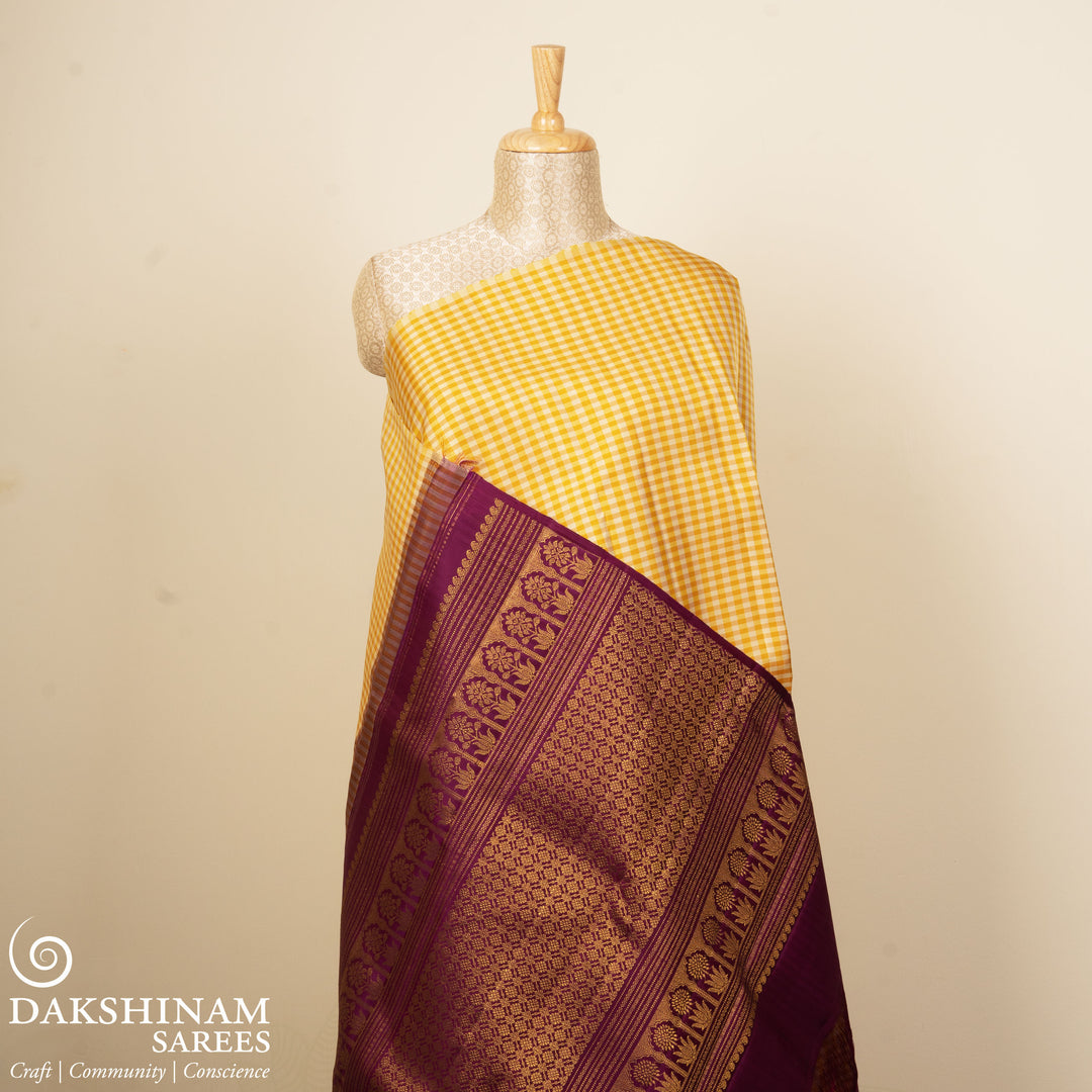 Beige and yellow small checked Kanjivaram silk saree with purple pallu featuring floral motifs, chevron and chakram designs, paired with dotted butta blouse.