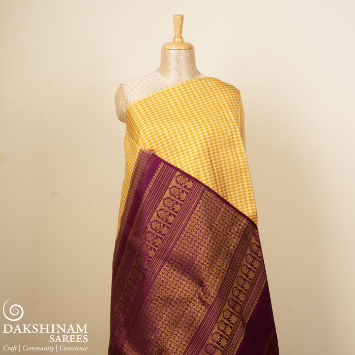 Beige and yellow small checked Kanjivaram silk saree with purple pallu featuring floral motifs, chevron and chakram designs, paired with dotted butta blouse.